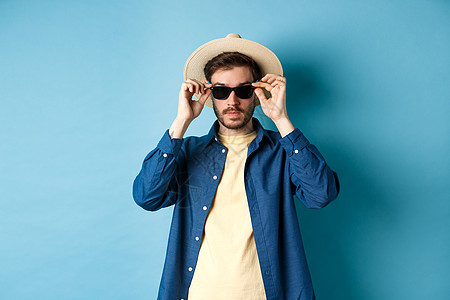 穿着草帽的英俊游客 在暑假时戴上墨镜 站在蓝背景上眼镜男性胡子旅游成功帽子办公室太阳镜成人胡须图片