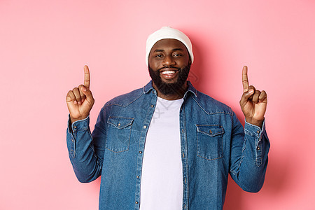 身穿时装和衣衫 粉红色背景的华丽英俊的非洲美国人男子举起手指 微笑并展示宣传性提议购物男人工作室促销横幅潮人胡须学生广告帽子图片