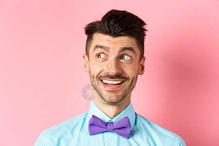 留着小胡子的逗趣微笑男人的特写 他指着并看起来很开心 盯着标志 站在粉红色的背景上快乐促销成人胡子情绪办公室发型领结成功商业图片