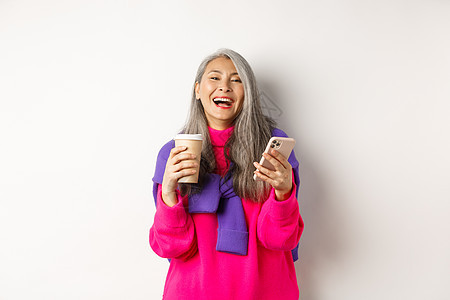 社交网络 快乐的亚洲年长妇女喝咖啡 拿着智能手机 嘲笑相机 站在白背景上站立 笑声派对技术喜悦老年母亲广告毛衣化妆品微笑乐趣图片