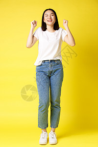 穿着牛仔裤和白色 T 恤的快乐亚洲女性的全尺寸照片从幸福中跳出来 赢得奖品和欢欣鼓舞 欢呼雀跃 欢呼雀跃 黄色背景理发黑发发型成图片