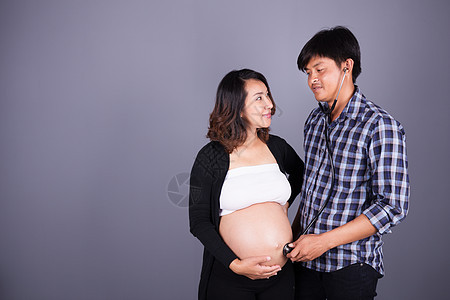 男人在灰色背景下用听诊器倾听其怀孕妻子的肚子丈夫父亲母性夫妻生活腹部女士女性成人男性图片