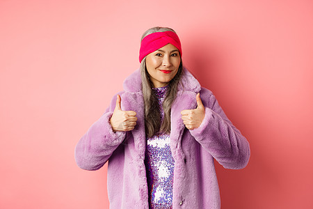 时装和购物概念 亚洲穿冬大衣的时装女士展示了拇指 微笑和看着满意 推荐公司宣传 粉红色背景等做法外套派对成人化妆品毛皮头发女性喜图片