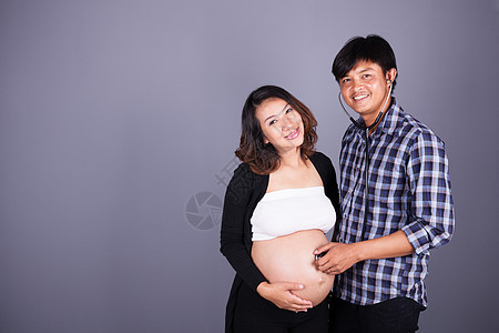 男人在灰色背景下用听诊器倾听其怀孕妻子的肚子女性腹部女士母亲父母婴儿夫妻成人男性丈夫图片