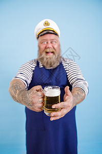 微笑的肥胖男人水手站在浅蓝色背景上 专注于用啤酒亲手图片
