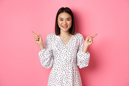 美丽的亚洲女性在购物上做出选择 指手指侧面和展示变体 对着镜头微笑 站在粉红背景的面前冒充理发青少年学生广告工作室黑发皮肤情感女图片