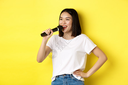 快乐的亚洲女人在卡拉 OK 里唱歌 拿着麦克风 带着欢快的微笑望向一旁 站在黄色背景上成人学生广告情感促销手势青少年黑发交易商业图片