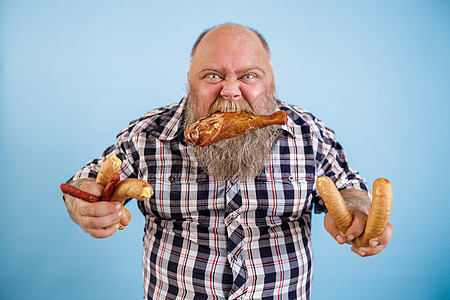 贪婪的肥胖男人把鸡腿塞在牙齿里 还有一堆蓝底香肠图片