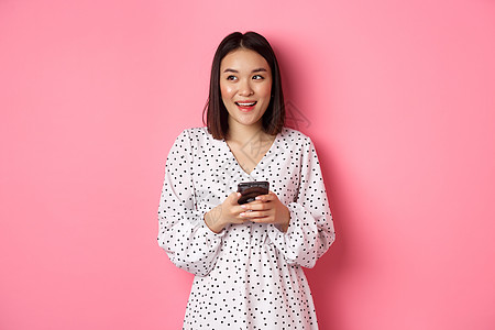 可爱的亚洲女性思考和微笑 在智能手机上发消息 浏览在线商店 站在粉红色背景上时看起来很梦幻横幅促销阅读女孩购物黑发思维技术工作室图片
