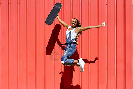 黑女人穿着随随便便的衣着 Wtih 一个滑板跳跃 快乐地跳在红色的城市墙壁背景街道编织幸福青年女孩黑发女性发型女士恐惧图片