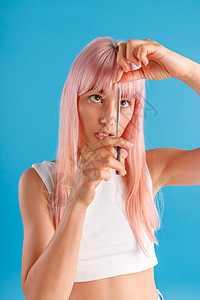穿着粉红色头发的可爱年轻女人 用剪刀剪短 站在蓝工作室背景上被孤立一旁图片