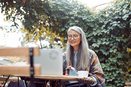 快乐的成熟亚洲妇女 在户外餐桌上用笔记本电脑前拿着眼镜和草莓甜点杯子食堂互联网桌子咖啡店成人女性手机商业城市图片