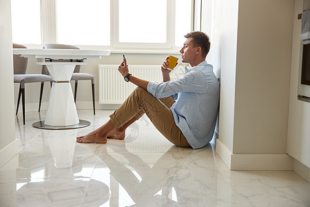 穿着蓝衬衫的白人年青男子坐在地板上 在现代厨房里寒冷椅子电话休息手机男人房间微笑快乐房子成人图片
