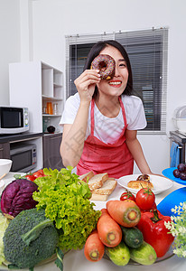 开心的女人在厨房房间 用巧克力甜甜圈遮着她的眼睛健康食物围裙小吃烹饪快乐饮食桌子女性甜点图片