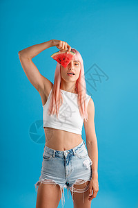 长着粉色头发的可爱年轻女人 在她眼睛上拿着一片西瓜 站在蓝色工作室背景旁与世隔绝图片