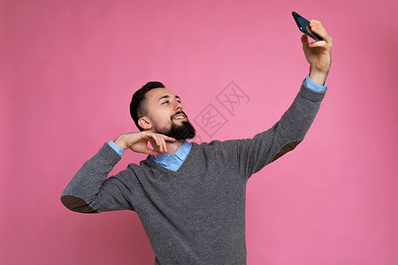 正面英俊的年轻黑发未剃须男子的照片 留着胡子 穿着休闲的灰色毛衣和蓝色衬衫 隔离在粉红色背景墙上 手持智能手机自拍照片 看着手机图片