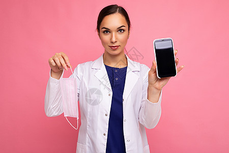 身着白色大衣 戴面罩的有吸引力的医疗年轻女青年妇女 显示现代移动电话在粉红背灌木上被隔绝 看着照相机图片