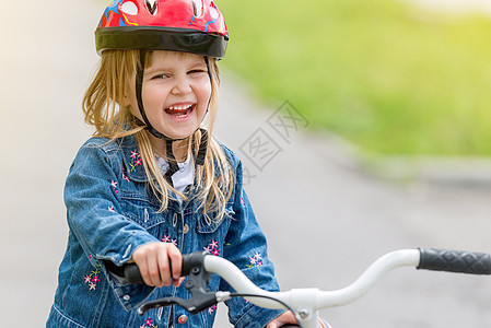 穿着头盔和牛仔衣的可爱小女孩 在自行车上蓝色红色夹克骑术喜悦帽子娱乐微笑牛仔布公园图片