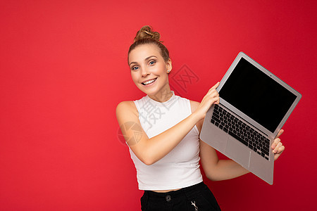 美丽微笑 迷人 快乐的金发年轻女子 头发聚集 看着相机 手持电脑笔记本电脑 身穿白色 T 恤 与红墙背景隔离互联网广告社交网页网背景图片