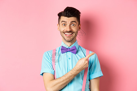英俊的caucasians男子与棕榈和胡子 指向右侧 展示广告 站在粉红背景上 在优雅的弓领情绪快乐发型成人促销成功办公室男人工图片