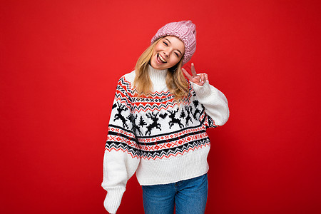 照片中 年轻积极快乐的金发美女带着真诚的情感 头戴粉色针织帽 身穿冬季套头衫 在红色背景中显得空荡荡的 展示着和平的姿态图片
