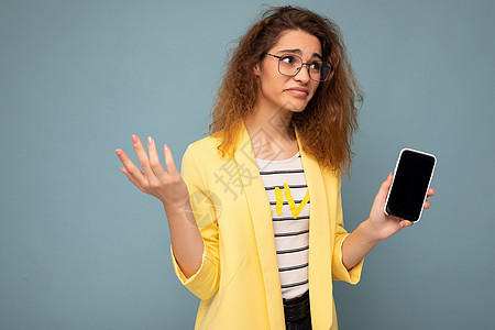 有魅力的年轻女性 一头卷曲的深金色头发 身穿黄色夹克 戴着光学眼镜 与背景隔离 手持并展示手机 手机上有空白空间 可以向侧面看 背景图片