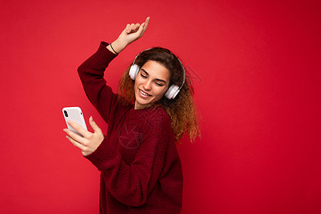 身穿深红色毛衣 头戴白色无线耳机 听音乐 使用手机上网 低头跳舞的美丽微笑的黑发卷发年轻女子被隔离在红色背景墙上的照片图片