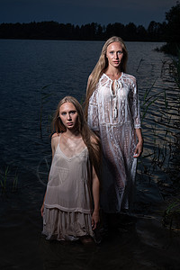 夏天晚上 两个年轻的双姐妹在湖水中穿着轻礼服头发双胞胎冒充女性海滩魅力池塘天空金发手电筒图片