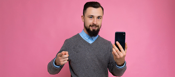 全景照片 帅气 酷酷 黑发 胡子拉碴的年轻男子留着胡须 穿着时尚的灰色毛衣和蓝色衬衫 站在粉色背景墙上 手持智能手机 用手机看着图片