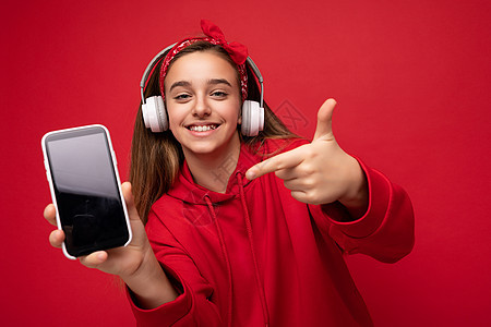 一张正面微笑的可爱黑发女孩身穿红色连帽衫的照片 与红色背景隔离 手持并展示智能手机 显示屏空空如也 戴着白色无线耳机 听着有趣的图片