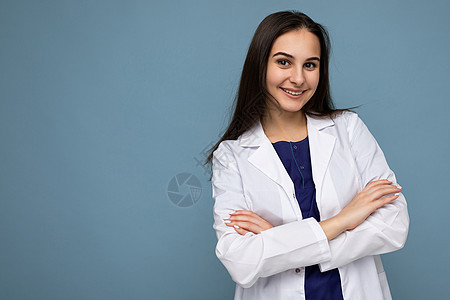 年轻漂亮 正面微笑的黑发女性的照片肖像 她身穿白色医用大衣 情绪真挚 与蓝色背景隔离 有复制空间 双臂交叉外套护士医师药品女士医图片