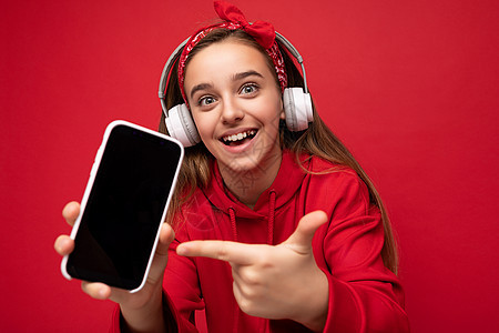 特写照片正面微笑惊讶可爱的黑发女孩穿着红色连帽衫隔离在红色背景持有和显示智能手机与空白显示用于切口戴着白色无线耳机听有趣的音乐看图片