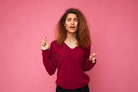 年轻迷人的黑发卷发女人的照片拍摄 她带着真诚的情感穿着时髦的粉红色毛衣 在粉红色的背景下与空旷的空间隔开 手指交叉祈求好运 手势图片