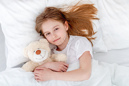 女孩和泰迪熊在床上童年微笑毯子就寝枕头拥抱白色苏醒卧室玩具图片