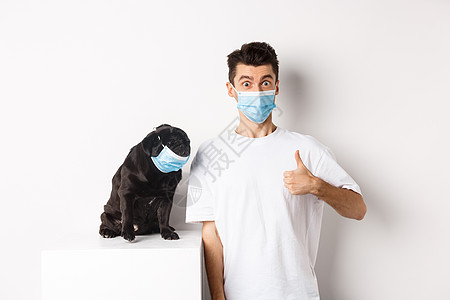 Covid19 动物和检疫概念 有趣的年轻人和小狗在医疗面具中的形象 主人举起拇指表示认可 赞美某些东西 白种背景促销微笑宠物乐图片