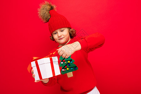 一个红墙背景卷发的酷男孩 穿着毛衣 手里拿着圣诞树 手里拿着礼盒购物情绪魅力盒子销售新年礼物红色背景图片