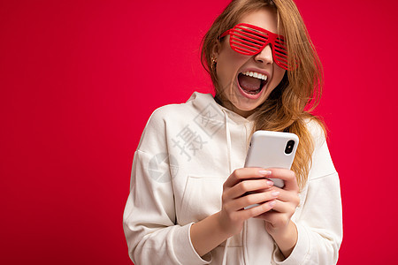 情绪激动的年轻金发女性穿着时尚的白色连帽衫 戴着滑稽的彩色眼镜 站在色彩缤纷的背景中 与世隔绝 手持和使用手机在互联网上在线交流图片