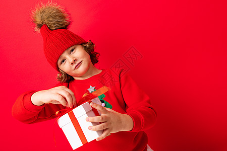 一个红墙背景卷发的酷男孩 穿着毛衣 手里拿着圣诞树 手里拿着礼盒情绪红色盒子新年礼物购物魅力销售背景图片