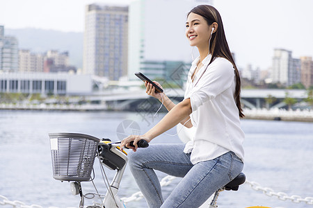 手持智能手机和在公园骑自行车的微笑女孩使用耳机运动技术城市生活女孩乐趣微笑女性音乐旅行街道图片