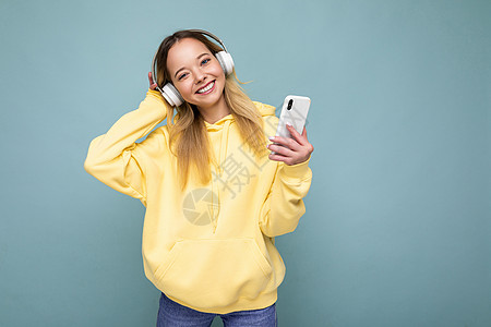 照片拍摄美丽快乐微笑的年轻女性穿着时尚休闲装隔离在色彩缤纷的背景墙上 戴着白色蓝牙无线耳机 听音乐 使用手机看着相机细胞收音机女图片