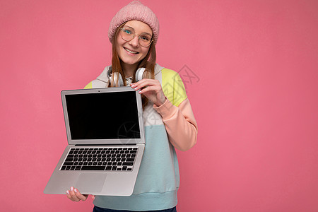 微笑的漂亮年轻女子的特写肖像 头戴彩色毛衣 戴着眼镜 手拿电脑笔记本电脑 上面有空白的复制空间屏幕监视器 头戴白色耳机 看着在粉图片