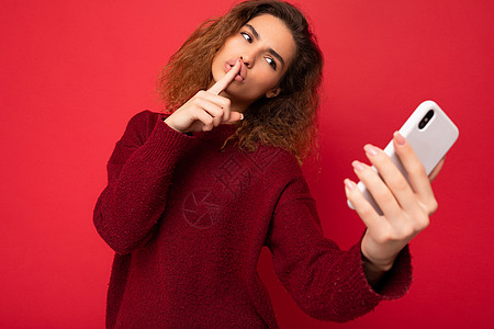 美丽的年轻女子 卷发 身穿深红色毛衣 与红色背景墙隔离 手持并使用智能手机 看着电话屏幕 自拍 表现出安静 保持秘密姿态图片