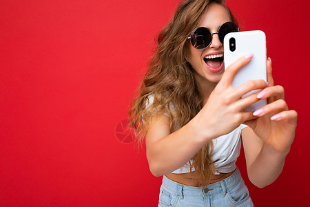 特写照片令人惊叹的快乐美丽的年轻金发女子手持手机使用智能手机相机自拍照片戴着太阳镜日常时尚服装隔离在彩色墙壁背景上看着设备屏幕图片