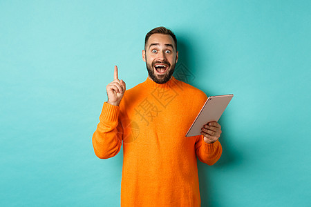 兴奋的成年男子使用数字平板电脑 并有一个想法 举起手指乌瑞卡标志 站在浅蓝色背景上药片促销胡子广告男人横幅胡须工作室橙子黑发图片