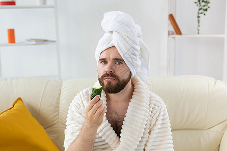 长胡子的可爱男人拿着黄瓜切片脸部 在家里 身体和皮肤上照顾男性概念美容产品护理面具奢华保湿奶油润肤治疗保健图片