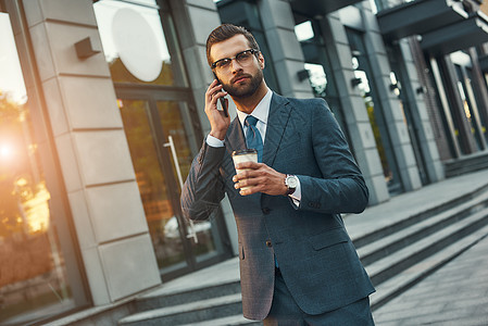 重要谈话 年轻和英俊的留胡子大胡子商务人士在户外站着时 用电话正式穿戴说话 喝咖啡和咖啡图片