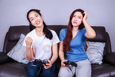 两个年轻女子在家沙发上玩电子游戏 使用棍棒控制器房子乐趣安慰友谊长椅快乐闲暇姐妹胜利朋友们图片