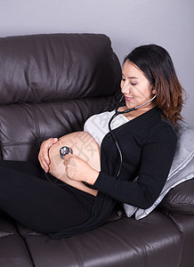 孕妇在腹部听婴儿的心跳并用听诊器检查其肚子妈妈白色沙发长椅说谎怀孕母亲成人医疗父母图片