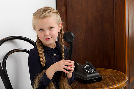 金发女孩用旧黑电话说话桌子讲话头发乐趣裙子相机女性青年椅子冒充图片