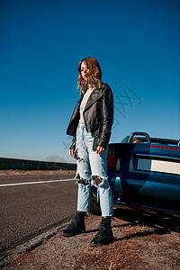 身穿皮夹克的无屋顶汽车附近的年轻妇女太阳敞篷车皮革海滨乐趣旅行女性陆地微笑蓝色图片